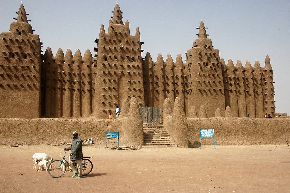 The Great Mosque of Djenné in daylight, with a man and his bicycle and goats in front of it.
