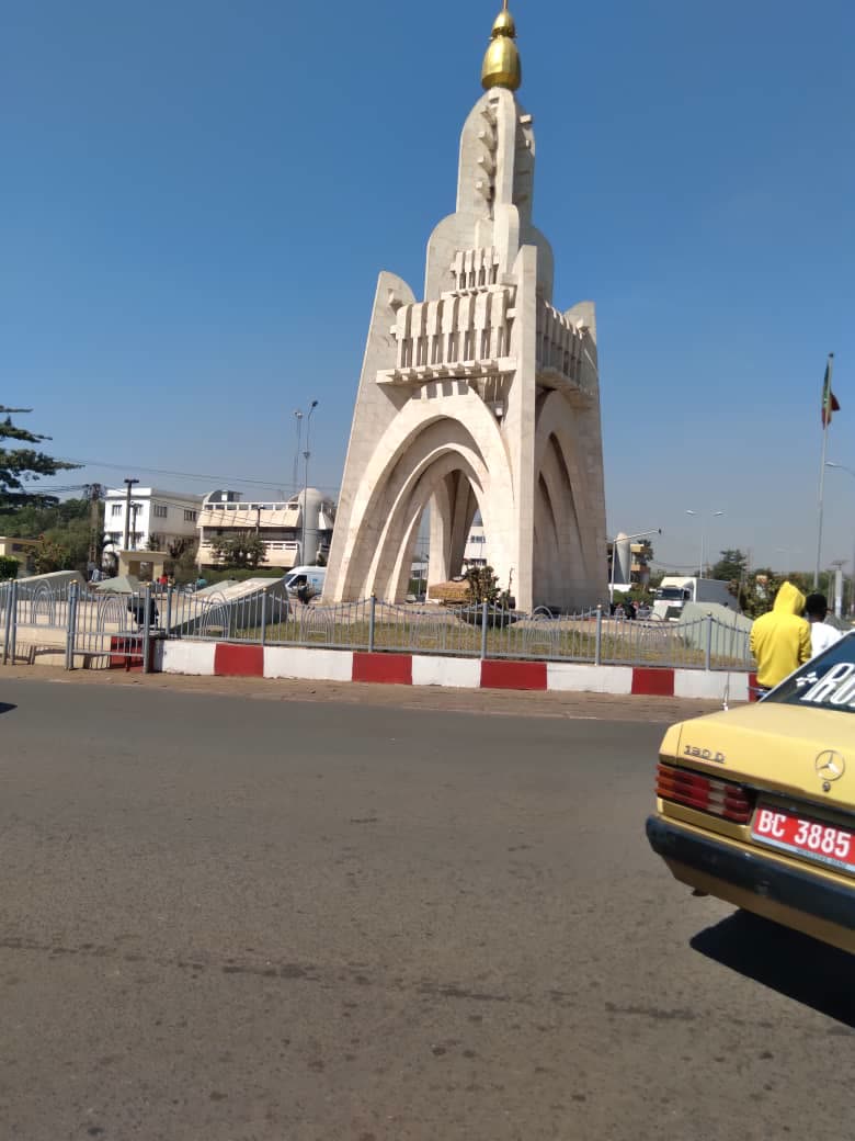 The Monument de l'Indépendance in Bamako under a blue sky, with a yellow taxi in the foreground.